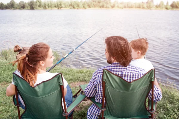 Una vista desde atrás. Los padres felices tienen a sus hijos de rodillas. Los adultos están sentados en sillas plegables y mirándose unos a otros. Los niños sostienen cañas de pescado en las manos . — Foto de Stock