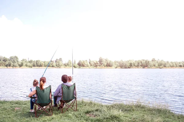 Gran vista del gran río que fluye. Hay una familia sentada junto a su orilla y disfrutando del momento. Están sosteniendo a los niños en el regazo. Los niños sostienen cañas de pescado en las manos . — Foto de Stock