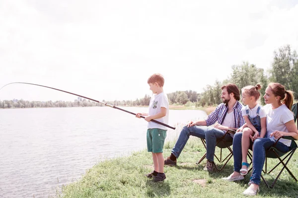 Garçon est debout au bord de la rivière et tenant la tige de poisson dans les mains. C'est vraiment long. Ses parents et sa sœur sont assis derrière eux et regardent . — Photo