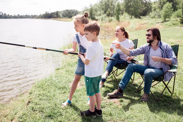 Niña y niño están sosteniendo largo caña de pescado juntos. Se concentran en catchinf algunos peces en el río. Sus padres están sentados detrás de ellos y cuidándolos. . — Foto de Stock