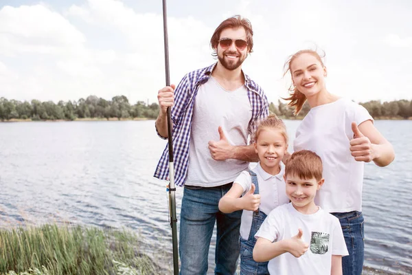 Portrait d'une famille heureuse debout ensemble et montrant le symbole similaire. Ils regardent droit sur la caméra et sourient. L'homme porte des lunettes et tient la tige de poisson dans la main droite . — Photo