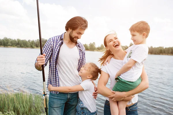 La familia feliz está unida. La mujer se ríe en voz alta y sostiene al hijo en sus manos. La niña está abrazando a su padre. El tipo sostiene una caña de pescado en la mano derecha. . — Foto de Stock