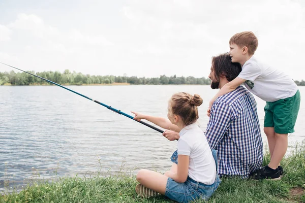 Papá y sus hijos están pasando tiempo juntos en la orilla del río. Están tratando de atrapar algunos peces. El hombre sostiene la caña de pescado en las manos. El chico está detrás de él y abrazándose. Chica está apuntando hacia adelante . — Foto de Stock