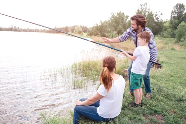 Guy apprend à son fils à pêcher correctement. Le garçon tient une longue tige de poisson. Le gars est le guider. La jeune femme est assise près d'eux . — Photo