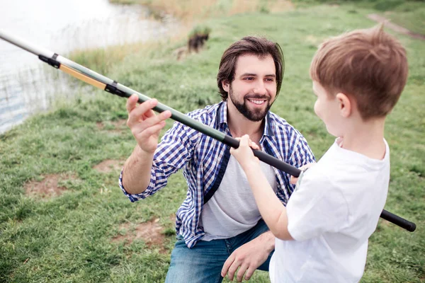 Happy dad is giving fish-rod to his son. He is smiling. Boy is holding fish-rod very tight and looking at dad.