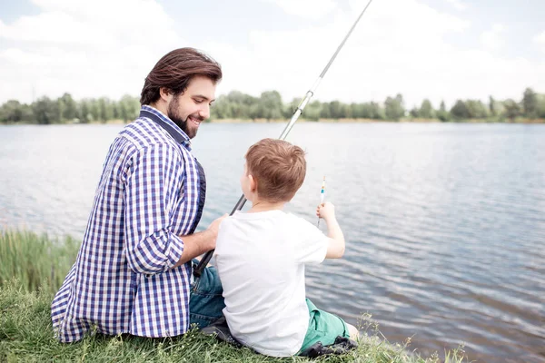 Bonita vista de feliz hijo y papá sentados juntos en la orilla del río. Guy está mirando a su hijo y pescando. Boy está mirando a su padre y hablando con él . — Foto de Stock