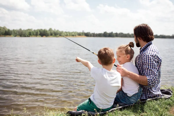 Obraz člověka a jeho děti seděli na břehu řeky. Guy je lov, zatímco jeho děti se dívají na něj. Muž drží dlouhou ryb tyč. — Stock fotografie