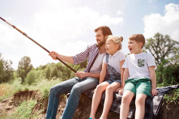 Una foto del hombre y sus hijos sentados juntos en la orilla del río. El tipo está pescando mientras sus hijos lo están viendo. El hombre sostiene larga caña de pescar . — Foto de Stock