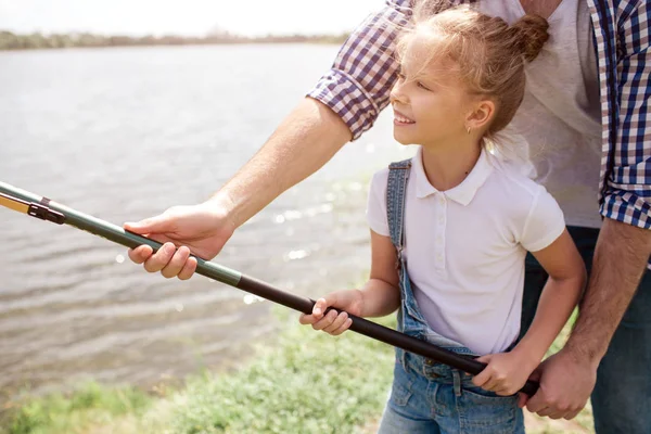 Una foto de un tipo ayudando a su hija a sostener la caña de pescar de una manera correcta. La chica lo sostiene con ambas manos y sonríe. Parece feliz. . — Foto de Stock