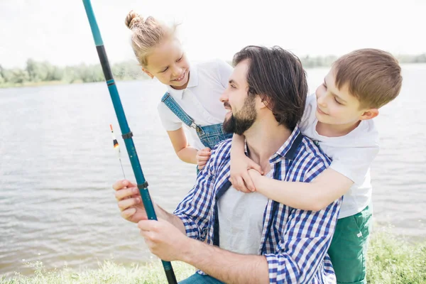 Los niños están detrás de su padre, abrazándolo y mirándolo mientras el hombre sostiene la caña de pescado y mira a su hija. Están parados en la orilla del río. Son felices. . — Foto de Stock