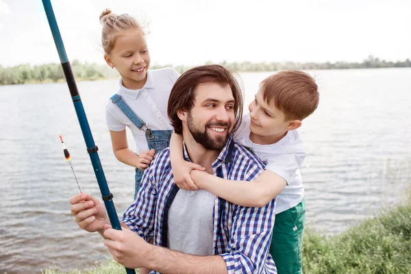 Una imagen de familia feliz de pie juntos en la orilla del río. El chico está abrazando a papá mientras el hombre lo mira y sonríe. También el tipo sostiene caña de pescado. Chica está de pie detrás de padre y mirándolo . — Foto de Stock