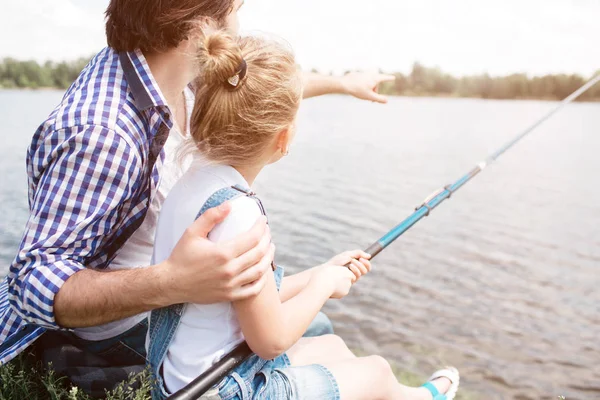 L'homme est assis avec sa fille au bord de la rivière et pointe vers l'avant. Fille tient la canne à poisson et la pêche . — Photo