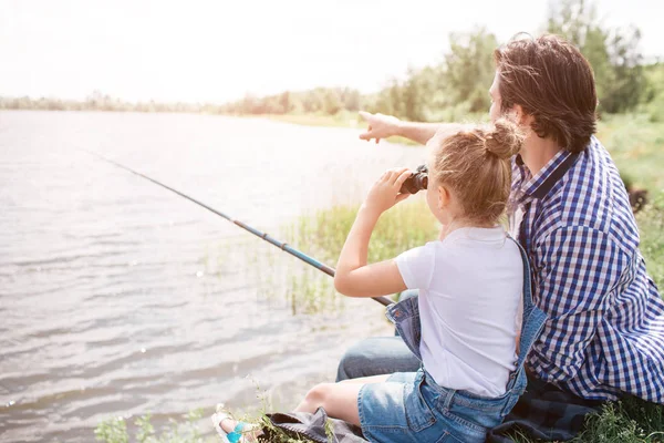 Adam suya yakın çimlerin üzerine kızıyla birlikte oturan ve ileriye dönük. Kız orada dürbün ile bakıyor. O balık-çubuk elinde tutan. — Stok fotoğraf
