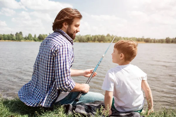 Otec a syn jsou seděli na trávě a při pohledu na sebe. Chlap drží ryby rod v rukou. On je rybaření. Vousatý muž se usmívá. — Stock fotografie