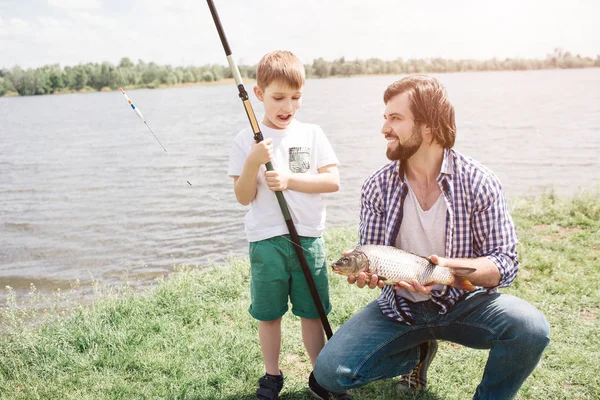 Padre e hijo están sentados juntos en la hierba y mirándose el uno al otro. El tipo tiene la caña de pescado en las manos. Está pescando. El hombre barbudo sonríe . — Foto de Stock