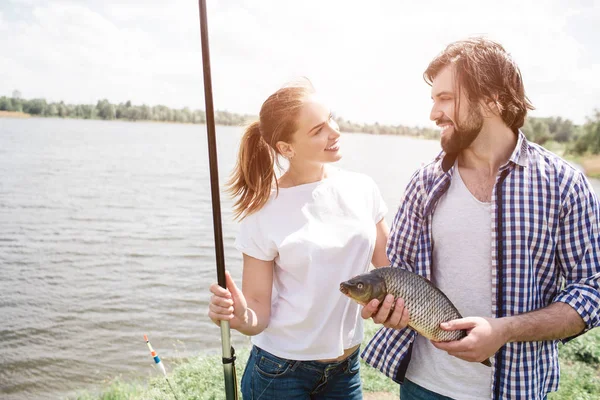 Belle image de couple debout ensemble au bord de la rivière et se regardant. Ils sourient. Le gars tient le poisson dans les mains tandis que la fille tient la tige de poisson . — Photo