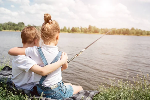 De magnifiques enfants sont assis ensemble près du lac et se serrent dans leurs bras. La fille pêche. Elle tient une canne à poisson. Ils sont seuls là-bas . — Photo