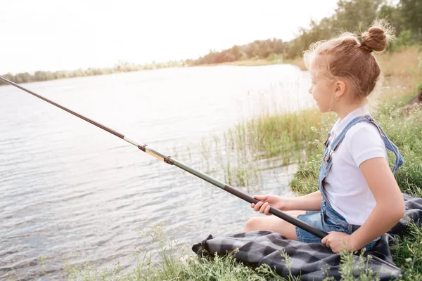 En bild av flicka sitter ensam på floden shore. Hon är fiske. Flickan håller fisk-stång med båda händerna. Hon tittar på vatten. Flickan är mycket allvarliga. — Stockfoto