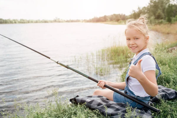 Flickan sitter på filt ensam och håller fisk-rod i höger hand. Hon är också tittar på kameran och leende. Flickan är att hålla sin bitars tumme. — Stockfoto