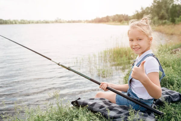 La chica está sentada en la manta sola y sosteniendo la caña de pescado en la mano derecha. También ella está mirando a la cámara y sonriendo. Chica está manteniendo su pulgar hacia arriba . — Foto de Stock