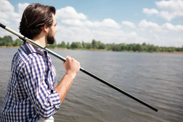 Tranquilo y reflexivo tipo está de pie a la orilla del lago y mirándolo. Él sostiene la caña de pescado en su hombro derecho con la mano derecha. Hay un tiempo hermoso outisde . — Foto de Stock