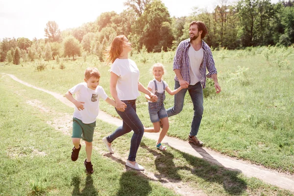 Obrázek celé rodiny chůze a skákání společně na cestách. Jdou přes zelenou louku. Smějí se. — Stock fotografie