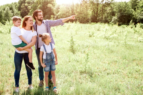 Bonita foto de la familia de pie juntos en el prado. Mamá sostiene a su hijo en las manos. La chica está parada junto a sus padres. Guy está apuntando más abajo. . — Foto de Stock