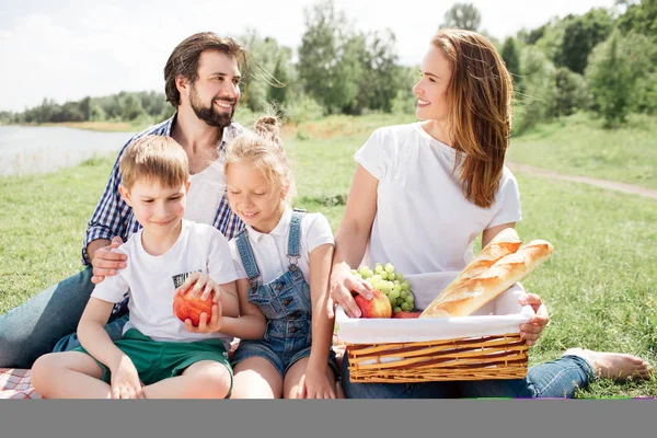 Los padres están sentados en una manta con sus hijos. La mujer sostiene una canasta con comida. Ella está mirando al hombre. Él le sonríe. Los niños mantienen unida una manzana . — Foto de Stock