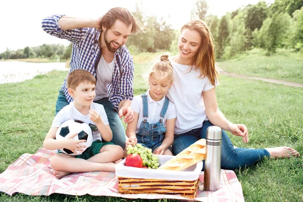 La familia está sentada y mirando la comida en la cesta. Quieren comerlo. Todos sonríen. Niño sostiene un fútbol en las manos . — Foto de Stock