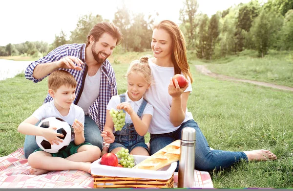 Hladová rodina sedí na dece a pohledu na košík s jídlem. Žena má apple ve svých rukou. Dívka je v rukou drží hrozny. Chlapec jí část hroznů. Muž chce chytit potravin z košíku. — Stock fotografie