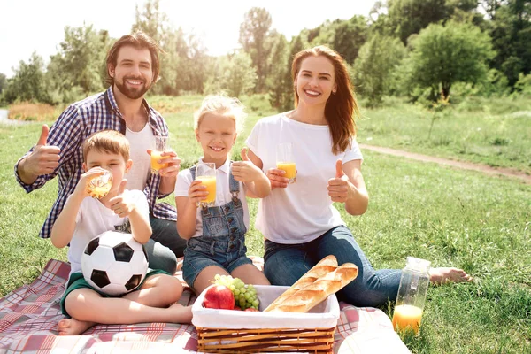 La familia alegre está sentada en una manta y mirando a la cámara. Están sonriendo. Cada uno de ellos tiene un vaso con zumo de naranja. Están mostrando el símbolo similar . — Foto de Stock