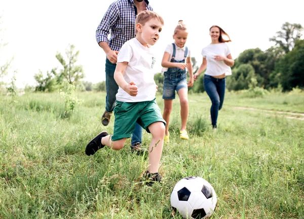 Una foto de la familia jugando al fútbol juntos. El chico corre delante de todos. Hay un hombre corriendo detrás de él y las niñas también. Están jugando en el prado . — Foto de Stock