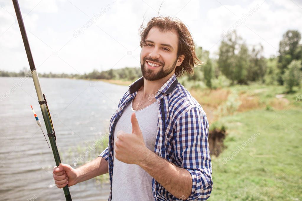 Calm and thoughtful guy is standing at the edge of lake and looking at it. He is holding fish-rod on his right shoulder with right hand. There is a beautiful weather outisde.