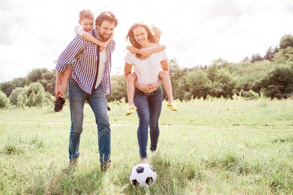 Parent are walking down the meaadow and holding kids on their back. Woman is playing with ball on grass with her leg. man and woman are looking at ball.