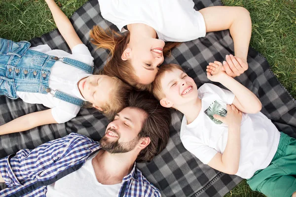 Hermosos miembros de la familia yacen en la manta sobre la hierba. El hombre está mirando a su hija. La mujer está mirando al hijo. Sonríen y ríen. La familia está cultivando tiempo juntos . — Foto de Stock