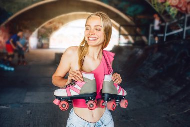 Sarışın kız gülümsüyor. Rollerbladers için bir eğitim odada duruyor. Silindirler boynunu üzerinde asılı olan. O onları ile elele geziyor. Kız gülümseyerek ve sola arıyorsunuz.