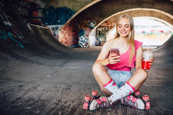 Smiley-Mädchen sitzt mit überkreuzten Beinen auf dem Boden und schaut auf das Telefon. Sie hält es in der rechten Hand und hält einen Plastikbecher mit Saft in der linken. Mädchen macht nach Sport Pause. — Stockfoto