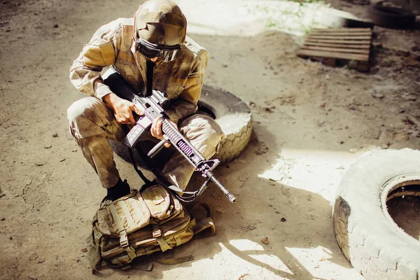 A picture of soldier sitting on the ground and wearing face mask. He is holding black rifle in hands. Man is looking odwn to bag. He has some rest.