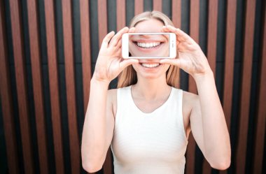 Bir telefon ile yüzünü kaplayan kızın komik resmi nerede gülümseme bir resmi vardır. Kız de gülümsüyor. Çizgili arka plan üzerinde izole.