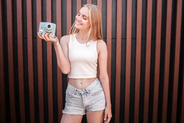 Happy blonde girl is standing at wall and holding blue camera in hands. She is looking on it and smiling. Isolated on striped background. — Stock Photo, Image