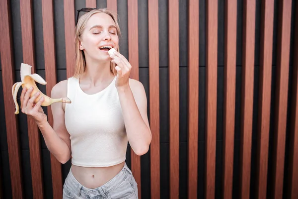 Mädchen hält Banane in der einen Hand und ein Stück davon in der anderen. Sie wird in diesen Frieden beißen. Mädchen genießt den Moment. isoliert auf gestreiftem Hintergrund. — Stockfoto