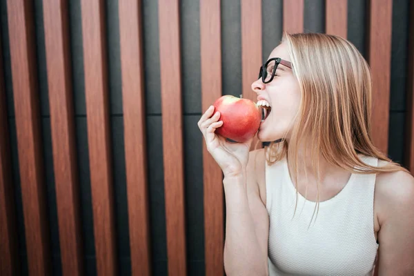 Ein Bild des blonden Mädchens, das in einen roten Apfel beißt. Sie genießt den Moment. isoliert auf gestreiftem Hintergrund. — Stockfoto