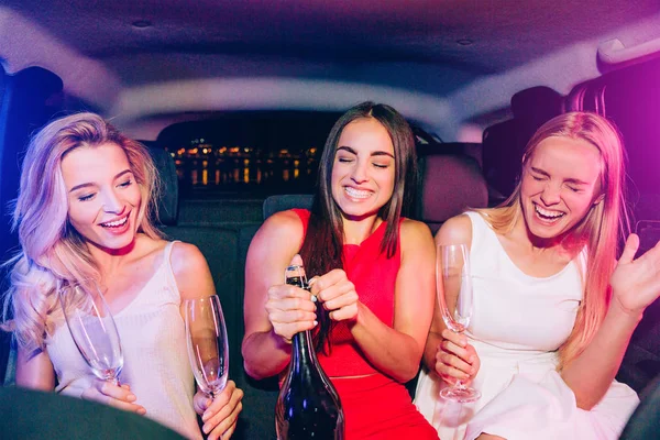 Schöne Mädchen sitzen im Auto. Brünette öffnet Flasche mit Alkohol. Die blonden Mädchen sind darüber sehr aufgeregt. junge Frauen halten Sektgläser in der Hand. — Stockfoto