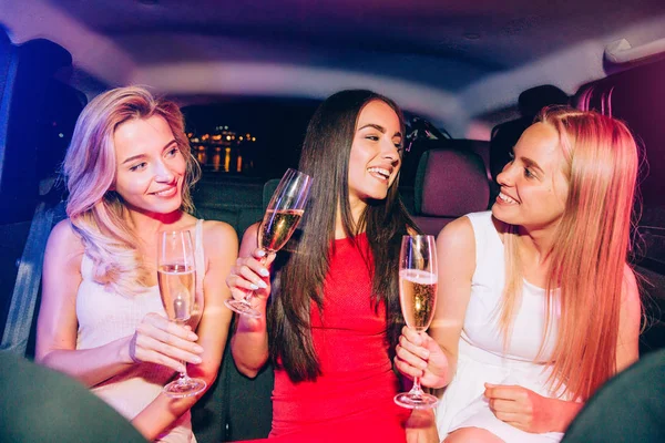 Positie and gorgeous girls are sitting in car and having fun. They are holding glasses with champagne. Girls are looking at each other and smiling. — Stock Photo, Image