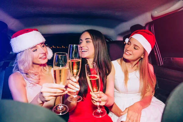 Blonda tjejer bär jul hattar. De tittar på deras brunett vän. De är glada och ler. Unga kvinnor har glas champagne i handen. — Stockfoto