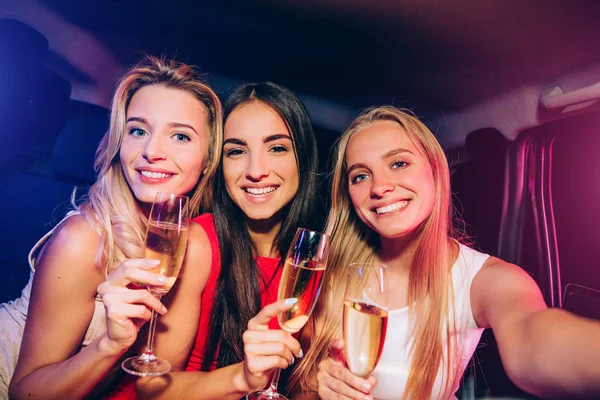 Mädchen sitzen im Auto und schauen geradeaus und lächeln. Mädchen auf der rechten Seite hält Kamera in der Hand. Alle haben Gläser mit Champagner. — Stockfoto