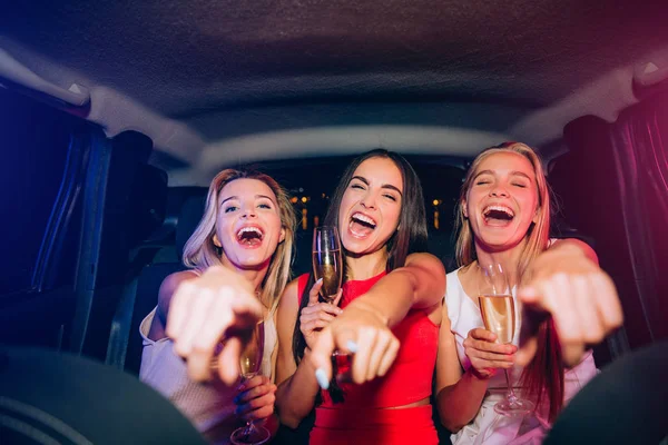 Glückliche und aufgeregte Mädchen sitzen zusammen im Auto. Sie schauen und zeigen auf die Kamera. Die jungen Frauen lachen und lächeln. Einige von ihnen haben ein Glas Champagner in der Hand. — Stockfoto