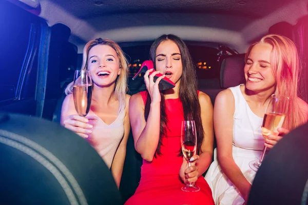Betrunkene Mädchen sitzen im Auto und trinken Champagner. Brünette spricht auf ihrem Schuh und täuscht ihr Handy vor. Sie hält die Augen geschlossen. Blonde Mädchen lachen laut. — Stockfoto