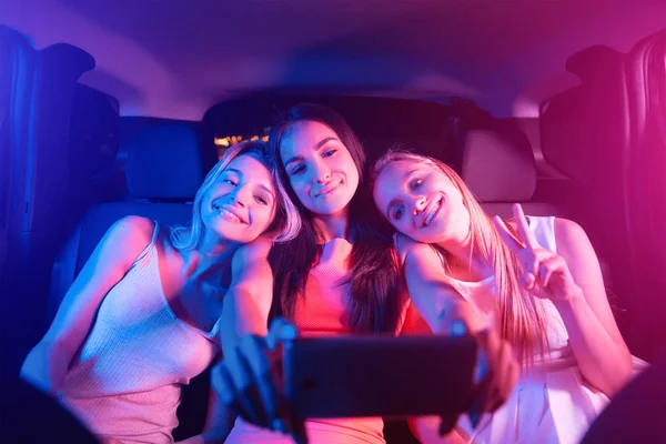Trevlig och glad tjej sitter i bil tillsammans. Blonda tjejer vilar på sina vänner axlar. Brunett håller svart telefon. Hon tar selfie. Unga kvinnor tittar på telefon. — Stockfoto