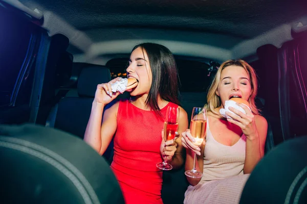 Hübsche Mädchen sitzen im Auto und essen Hamburger. Sie haben auch Sektgläser in der Hand. junge Frauen genießen ihre Mahlzeit. — Stockfoto